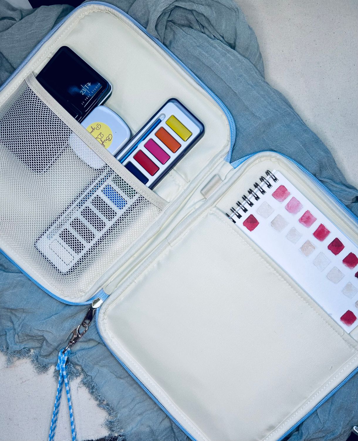 The Juliette Watercolor Clutch open, showing mesh pockets, a 23 Tree Studio paint set, brush, and watercolor swatch book inside